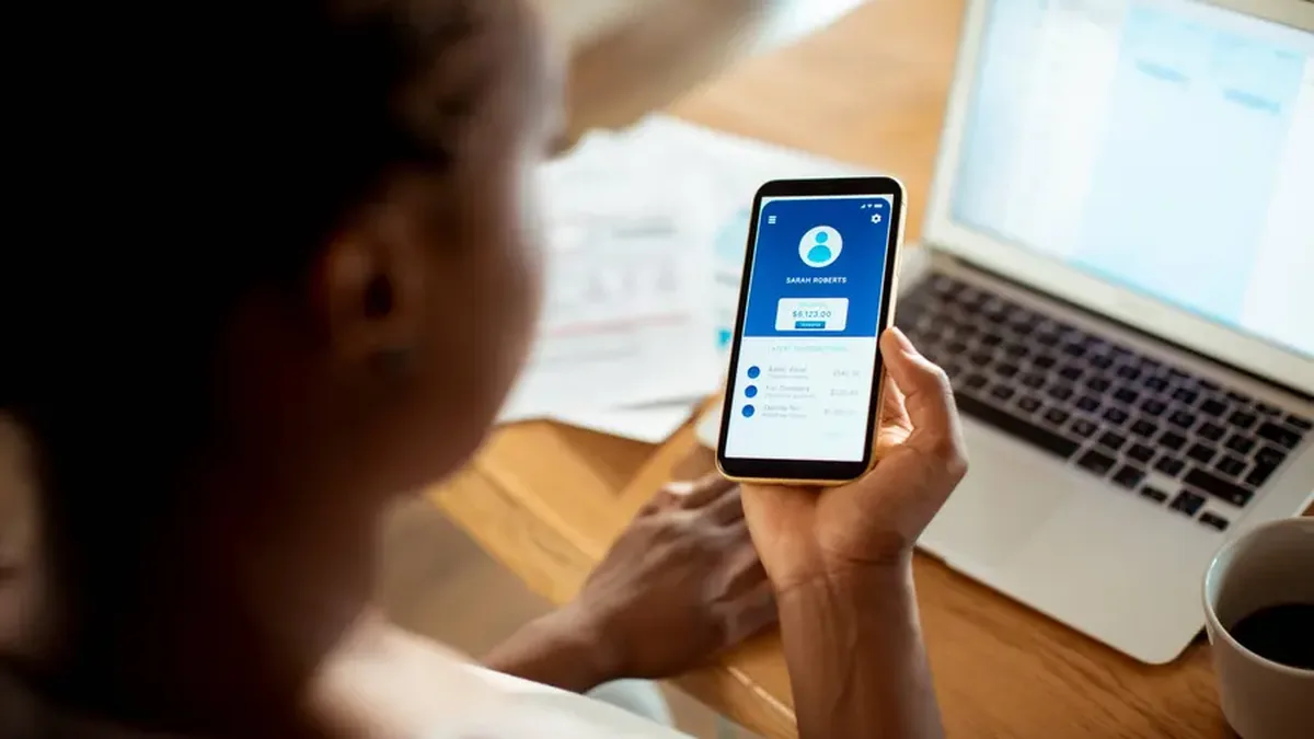 Une personne consulte son compte bancaire sur son téléphone, à côté d&rsquo;un ordinateur portable